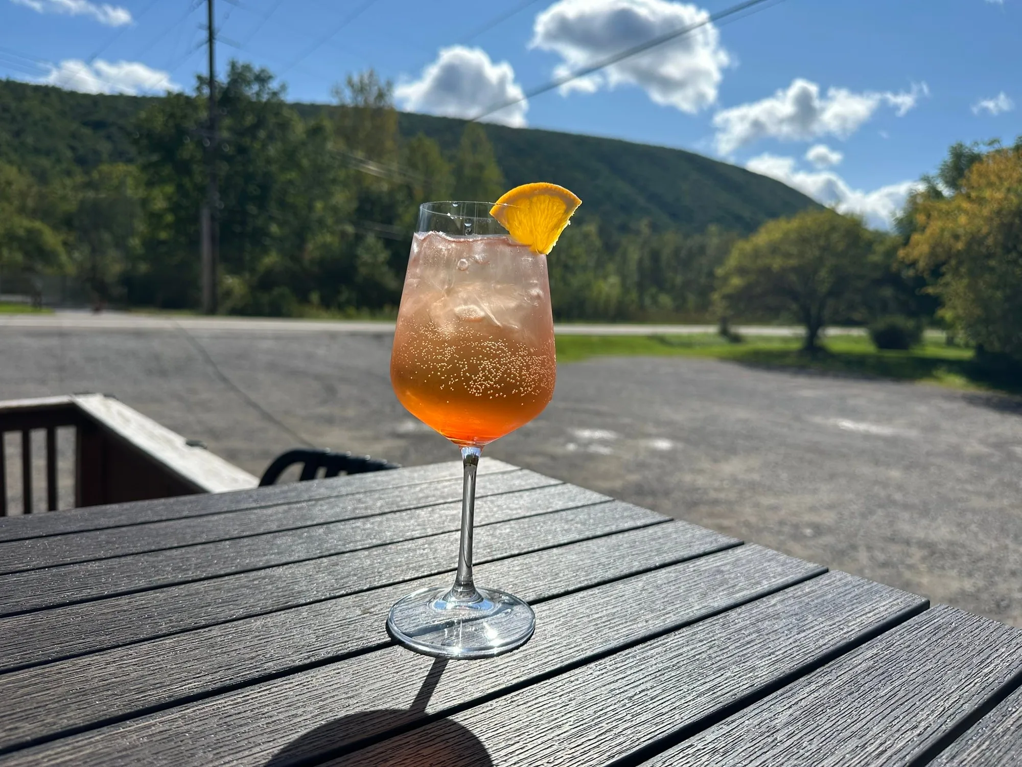 Aperol spritz with orange slice on an outdoor deck table, green hills and blue sky behind